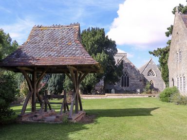 An outdoor lawn with a wooden structure with a tiled roof and stone buildings with arched windows in the background at The Alms House East in Forthampton near Tewkesbury