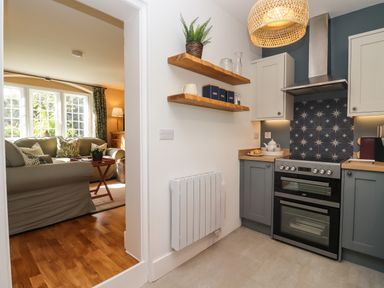A kitchen with grey cabinets a stove wooden shelves and a view into a living room with a sofa at The Alms House East in Forthampton near Tewkesbury