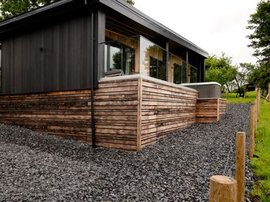 A modern house with black and wooden exterior and glass railings surrounded by gravel and trees at The Lookout in Longridge