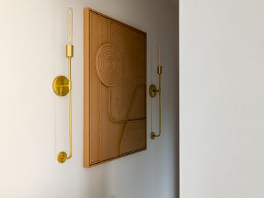 A wall with a textured abstract art piece and two vertical gold wall sconces with exposed bulbs at The Lookout in Longridge