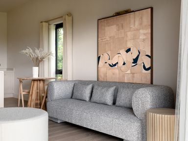 A living room with a gray sofa three cushions an abstract wall painting a wooden round table with chairs a vase with flowers and a round ottoman at The Lookout in Longridge