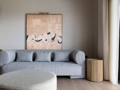 A living room with a gray sofa three cushions a round ottoman a wooden side table and abstract wall art at The Lookout in Longridge