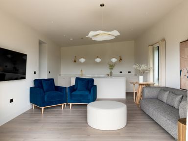 A living room with two blue armchairs a round white ottoman a grey sofa a small wooden table with a vase and flowers a wall-mounted TV and a kitchen area in the background at The Lookout in Longridge