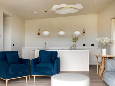 A kitchen area with white cabinets gold fixtures blue armchairs a wooden round table with flowers and a gray ottoman at The Lookout in Longridge
