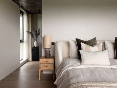 A bedroom with a bed with multiple pillows a wooden nightstand with a lamp and a vase on a pedestal near a window at The Lookout in Longridge