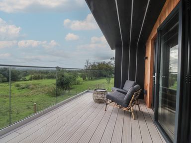 A balcony with glass railing a wicker chair with cushions and a side table overlooking a green field at The Lookout in Longridge
