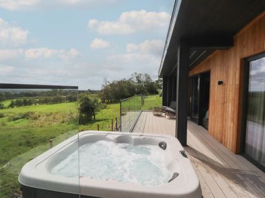 An outdoor deck with a hot tub glass railing and seating area overlooking green fields at The Lookout in Longridge