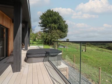 An outdoor wooden deck with a covered hot tub and glass railing overlooking a green field at The Lookout in Longridge