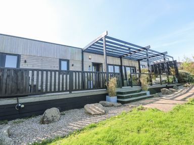 A wooden terrace with glass roof and stairs leading to a pathway with rocks and grass at Bay View in Portreath