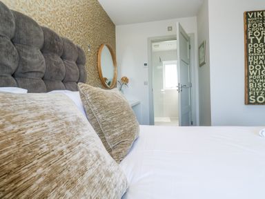 A bedroom with a bed having brown pillows and a gray padded headboard a round mirror on the wall and an open door leading to a bathroom at Bay View in Portreath