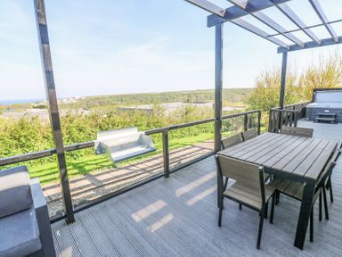 A patio with a dining table and chairs a barbecue grill and a hot tub overlooking greenery and hills at Bay View in Portreath