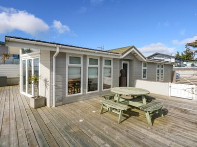 An outdoor wooden deck with a round picnic table and benches next to a building with multiple windows and a glass door at The Warren M45 in Abersoch