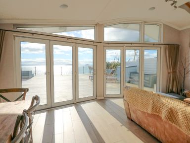 A living area with wooden dining chairs a sofa and large glass doors opening to a deck with ocean view at The Warren M45 in Abersoch
