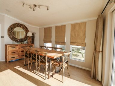 A dining room with a wooden table and chairs next to windows with blinds and a sideboard with a round mirror at The Warren M45 in Abersoch