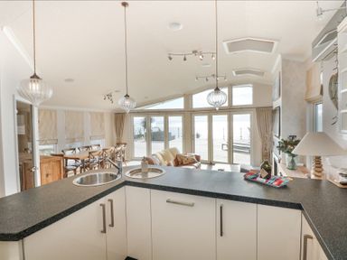 An open kitchen and dining area with a black countertop island, dining table, and large windows overlooking a deck outside at The Warren M45 in Abersoch