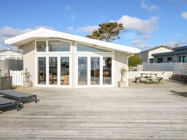 A house with large glass doors and windows on a wooden deck with lounge chairs and a picnic table at The Warren M45 in Abersoch