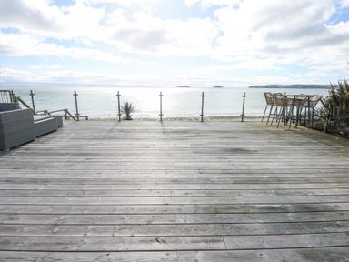 A wooden deck with glass railing overlooking a beach and ocean with outdoor seating at The Warren M45 in Abersoch