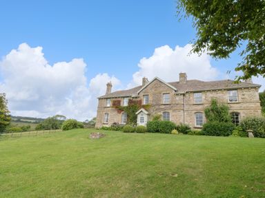 A stone house with chimneys surrounded by shrubs and grass under a blue sky at Ivy Cottage in Colerne