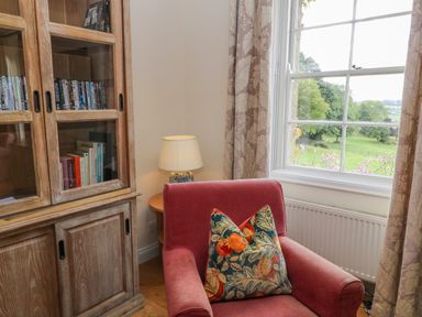 A red armchair with a patterned cushion next to a wooden cabinet with books and DVDs and a table lamp by a window with floral curtains at Ivy Cottage in Colerne