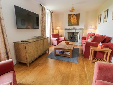 A living room with a wall-mounted TV wooden cabinet red upholstered chairs and sofa a wooden coffee table on a rug a fireplace with a wood stove and lamps at Ivy Cottage in Colerne