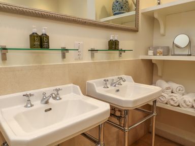 A bathroom with two white sinks glass shelves with bottles a mirror and rolled towels on shelves at Ivy Cottage in Colerne