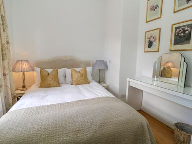 A bedroom with a bed with pillows and lamps on side tables a white dressing table with a mirror and floral pictures on the wall at Ivy Cottage in Colerne