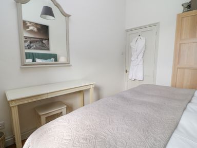 A bedroom with a bed covered by a patterned blanket a dressing table with a stool a large mirror on the wall a robe hanging on the door and a wooden wardrobe at Ivy Cottage in Colerne