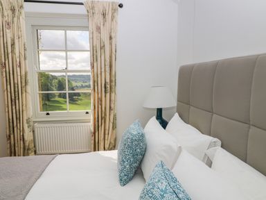 A bedroom with a bed featuring white sheets and blue patterned pillows a grey padded headboard a bedside lamp floral curtains and a window showing a green outdoor landscape at Ivy Cottage in Colerne