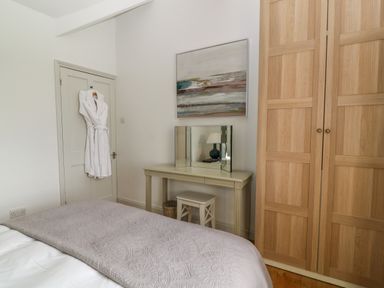 A bedroom with a bed, dressing table with mirror, stool, wardrobe, and a bathrobe hanging on the door at Ivy Cottage in Colerne