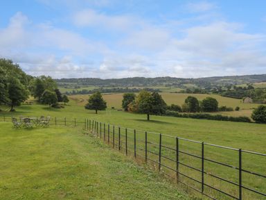 A grassy field with a metal fence and outdoor table and chairs overlooking rolling hills and trees at Ivy Cottage in Colerne
