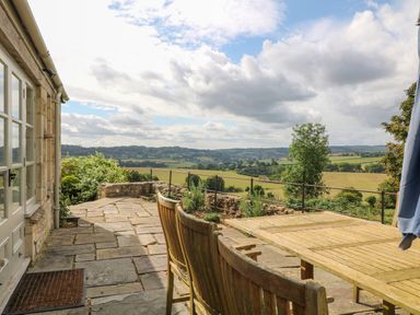 A stone patio with wooden chairs and a table overlooking fields and hills at Ivy Cottage in Colerne
