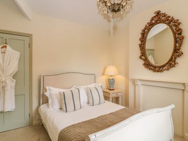 A bedroom with a bed pillows a lamp on a bedside table a hanging robe and ornate mirror at Peartree in Colerne