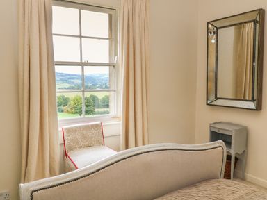 A bedroom with a bed curtain drapes an upholstered chair a small bedside table and a wall mirror at Peartree in Colerne