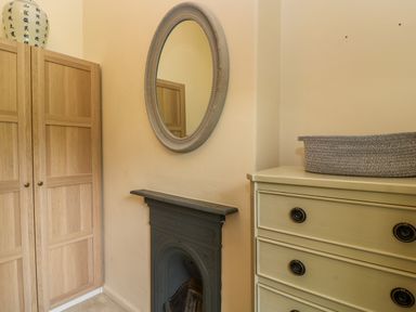 A corner of a room with a wooden wardrobe a round mirror above a black fireplace and a cream chest of drawers with a woven basket on top at Peartree in Colerne