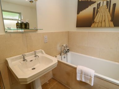 A bathroom with a white sink and bathtub with a towel hanging on the side and toiletries on a glass shelf at Peartree in Colerne