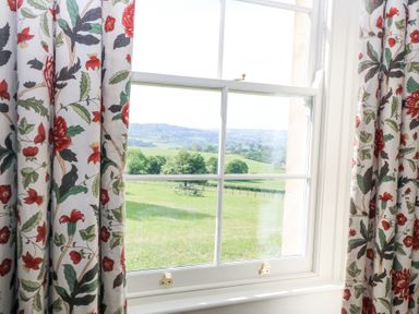 A window with floral curtains looking out to a green field with distant hills at Peartree in Colerne