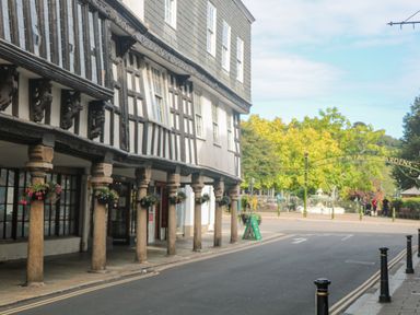 A street with a historic building supported by stone columns and hanging flower pots near a garden entrance gate