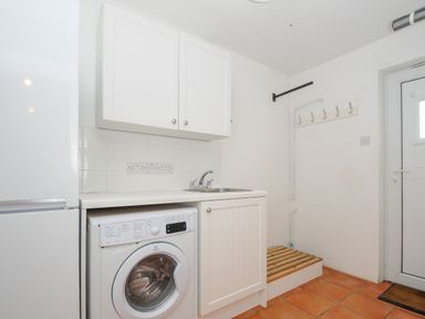 A utility room with a washing machine a sink white cabinets hooks on the wall and a door at Bryher in Trebetherick near Polzeath