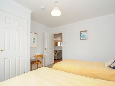 A bedroom with two beds covered in yellow bedspreads a wooden chair and desk a door leading to a living room a white closet and a picture on the wall at Bryher in Trebetherick near Polzeath