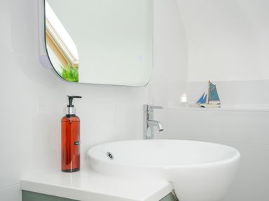 A bathroom countertop with a white sink a red soap dispenser a mirror and a small model sailboat at Bryher in Trebetherick near Polzeath