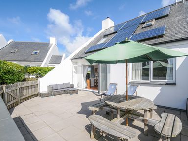 An outdoor patio with a wooden table and benches under a green umbrella two chairs a gated fence and a house with solar panels on the roof at Bryher in Trebetherick near Polzeath