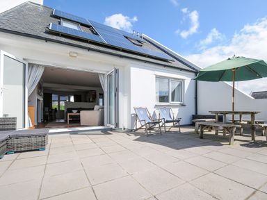 An outdoor patio with two wooden deck chairs a wooden picnic table with umbrella and sofa visible through open glass doors at Bryher in Trebetherick near Polzeath