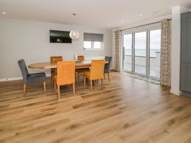 A dining room with wooden table and six chairs with gray and yellow upholstery and sliding glass doors with ocean view at Melin Y Plas in Llanaber