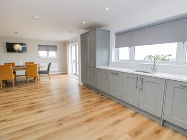 A kitchen and dining area with grey cabinets, a sink, wooden dining table with mustard and grey chairs and sliding glass door at Melin Y Plas in Llanaber