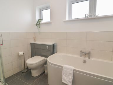 A bathroom with a toilet, bathtub with towel, two windows, and a grey storage unit at Melin Y Plas in Llanaber