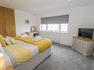 A bedroom with a bed with yellow bedding a dresser with a mirror a chest of drawers with a television and a window with a grey blind at Melin Y Plas in Llanaber