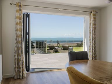 A dining area with a round wooden table and chairs overlooking a patio with lounge chairs and an ocean view at Melin Y Plas in Llanaber