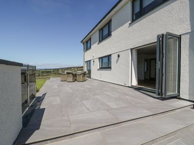 A large tiled patio with a wicker table and chairs outside a two-story white house with black framed windows at Melin Y Plas in Llanaber