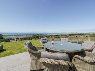 An outdoor patio with a glass table surrounded by six cushioned wicker chairs overlooking a grassy area and the ocean at Melin Y Plas in Llanaber