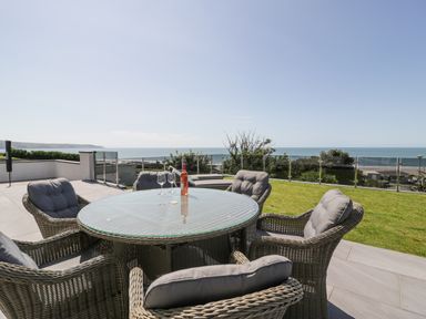 An outdoor patio area with a round glass table surrounded by six wicker chairs with cushions and a bottle of wine and two wine glasses on the table by Melin Y Plas in Llanaber